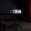 Na zdjęciu znajduje się biały projektor z napisem "4K" umieszczony na czarnym stoliku na kółkach, w ciemnym pomieszczeniu z zasłonami i sprzętem audio w tle. Projektor emituje jasne światło z obiektywu.