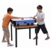 Na zdjęciu znajduje się stół do cymbergaja z niebieską powierzchnią do gry, ustawiony na prostym stole z metalowymi nogami. Na boku stołu widnieje napis "Air Hockey".