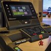 Na zdjęciu widoczny jest panel sterowania bieżni elektrycznej z dużym ekranem dotykowym, na którym wyświetlane są różne opcje treningowe oraz przyciski do regulacji nachylenia i prędkości. Na panelu znajduje się logo "BH" oraz przyciski "STOP" i "START" z
