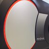 Zbliżenie na fragment roweru magnetycznego z widocznym okrągłym elementem w kolorze srebrnym, otoczonym czerwonym pierścieniem. Obok znajduje się perforowany panel z napisem "RHONE".