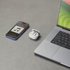 Na zdjęciu widoczne są białe słuchawki douszne w otwartym etui, leżące obok smartfona z wyświetlonym ekranem odtwarzacza muzyki oraz laptopa z widoczną klawiaturą. Na ekranie smartfona widnieje godzina 12:50 oraz ikona nuty.