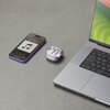 Na zdjęciu znajduje się otwarte etui z fioletowymi słuchawkami dousznymi, obok leży smartfon z wyświetlaczem pokazującym odtwarzacz muzyki z ikoną nuty i godziną 12:50, a także fragment laptopa z widoczną klawiaturą.