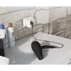 Na zdjęciu widać ładowarkę sieciową SBS Multi-Socket Charger 20W w kolorze białym podłączoną do gniazdka na tle ściany z kamienia. Obok znajdują się butelki z szamponem i odżywką oraz suszarka do włosów leżąca na blacie.