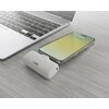 Na zdjęciu znajduje się biały powerbank z logo "SBS" obok smartfona z widoczną ikoną ładowania baterii oraz fragment laptopa na jasnym, drewnianym blacie. Powerbank ma zaokrąglone krawędzie i niewielkie diody sygnalizacyjne.