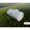 Biały powerbank z logo "sbs" leży na trawie, z widocznym portem USB i wskaźnikiem LED. W prawym dolnym rogu znajduje się napis "ECO Friendly RECYCLED MATERIAL".