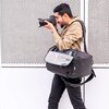Ciemnoszary plecak fotograficzny z otwartą komorą, w której znajduje się aparat z dużym obiektywem. Plecak ma wyściełane wnętrze i siatkową kieszeń na klapie.