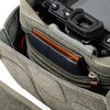 Szaro-zielona torba fotograficzna z widocznym wnętrzem, w którym znajduje się aparat fotograficzny oraz kieszeń z brązowym portfelem. Torba ma zamek błyskawiczny i wykonana jest z materiału o teksturze przypominającej tkaninę.