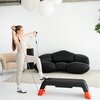 Na zdjęciu znajduje się czarno-pomarańczowy step z funkcją ławki HMS AS009 PRO, na którym stoi osoba trzymająca gumy oporowe. W tle widoczna jest czarna sofa, jasny dywan oraz dekoracyjne lampy wiszące.