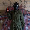 Osoba ubrana w zielony kombinezon i maskę ochronną trzyma w ręku telefon z wyświetlaczem, na którym widnieje napis "Motorola Moto G55". W tle znajduje się ściana pokryta graffiti w różnych kolorach.