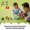 Na zdjęciu znajduje się zestaw klocków LEGO DUPLO z sorterem kształtów w kształcie zielonego pojemnika, wokół którego rozłożone są różnokolorowe klocki z motywami zwierząt i roślin. W górnej części zdjęcia widnieją symbole klocków z oznaczeniem "1½+" oraz