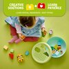 Na zdjęciu widoczny jest zestaw klocków LEGO DUPLO, w tym sorter kształtów w kształcie zielonej miski z pokrywką z otworami o różnych kształtach. W górnej części zdjęcia znajduje się napis: "CREATIVE SOLUTIONS + LEARN PATIENCE", a poniżej: "LEARN SPATIAL 
