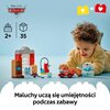 Na zdjęciu znajduje się zestaw klocków LEGO z serii DUPLO, przedstawiający warsztat z elementami takimi jak budynek, samochód i akcesoria warsztatowe. Widoczny jest również tekst: "Maluchy uczą się umiejętności podczas zabawy" oraz logo "Disney Pixar Cars