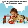 Na zdjęciu znajduje się zestaw klocków LEGO, przedstawiający warsztat z otwieranymi drzwiami oraz czerwony samochód. W tle widoczny jest napis: "Zygzak McQueen wjeżdża z impetem przez otwierane drzwi".