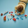 Na zdjęciu widać zestaw klocków LEGO DUPLO rozłożony na niebieskim tle, w tym czerwony samochód z napisem "Rust-eze" na masce oraz różne elementy konstrukcyjne, takie jak okno, klucz i inne akcesoria. Obok klocków znajduje się dziecko w jasnym ubraniu, kt