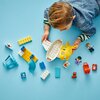 Na zdjęciu widać zestaw klocków LEGO DUPLO, w tym elementy samolotu, figurki postaci oraz różnokolorowe klocki na niebieskim tle. Widoczne są także akcesoria takie jak walizki i misiek.