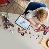 Na zdjęciu widać stół, na którym znajdują się klocki LEGO oraz tablet z wyświetlonym schematem budowy. Obok leży duży kilof zbudowany z różowych i brązowych klocków, a wokół rozłożone są różne elementy zestawu LEGO Minecraft.