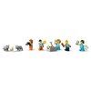 Na zdjęciu znajduje się zestaw klocków LEGO przedstawiający figurki ludzi w strojach zimowych oraz figurki zwierząt, w tym wilków i szczeniaka. W centrum znajduje się mały stół z akcesoriami, a jedna z figurek trzyma kamerę, inna pizzę, a kolejna mapę.