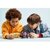Na zdjęciu widoczne są dwie osoby bawiące się zestawem klocków LEGO, przedstawiającym kolorowego robota z różowymi i niebieskimi elementami oraz figurkę w zielonym kolorze. Tło jest jednolite, w jasnym odcieniu, co podkreśla centralny element zdjęcia.