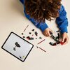 Na zdjęciu widać osobę w niebieskiej bluzie, która składa zestaw klocków LEGO, obok leży tablet z instrukcją budowy. Na stole rozłożone są różne elementy klocków, w tym czerwone i czarne części oraz miniaturowy miecz świetlny.