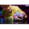 Na zdjęciu znajduje się zestaw LEGO przedstawiający kolorową figurkę przypominającą stworzenie z gry Fortnite, umieszczoną na ciemnym stole obok klawiatury i żółtych słuchawek. Obok figurki leży otwarta instrukcja z widocznym napisem "FORTNITE" oraz ilust