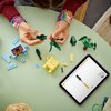 Na zdjęciu widać zestaw klocków LEGO, w tym elementy w kolorach zielonym, niebieskim i żółtym, ułożone na zielonym stole. Obok znajduje się tablet z instrukcją budowy, na której widoczny jest rysunek przedstawiający montaż elementów LEGO.