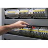 Na zdjęciu widać rękę trzymającą tester napięcia, który jest używany do sprawdzania instalacji elektrycznej w skrzynce z bezpiecznikami. Na testerze widoczny jest czerwony wskaźnik świetlny, a w tle znajdują się rzędy bezpieczników z żółtymi etykietami.