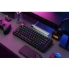 Na zdjęciu znajduje się klawiatura mechaniczna z podświetleniem RGB, otoczona różnymi akcesoriami elektronicznymi, w tym kontrolerem do gier, smartfonem, myszką i słuchawkami. Obok klawiatury widoczny jest cyfrowy zegar wyświetlający godzinę 13:88 oraz po