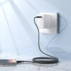 Na zdjęciu widać czarny kabel USB-C - USB-C podłączony do białej ładowarki w gniazdku ściennym, z drugim końcem podłączonym do smartfona leżącego na blacie. Kabel jest zwinięty w pętlę na powierzchni blatu, a w tle widać delikatne cienie na ścianie.