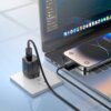 Na zdjęciu widać laptopa z otwartym ekranem, na którym wyświetlony jest interfejs graficzny, oraz smartfon leżący na klawiaturze laptopa. Kabel USB-C - USB-C VEN-DENS 240W 1.8m Czarny jest podłączony do smartfona i ładowarki w gniazdku elektrycznym; na wtyczce kabla widoczny jest napis "VEN-DENS".