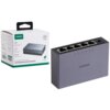 Na zdjęciu znajduje się szary switch UGREEN z pięcioma portami Ethernet, obok którego widoczne jest opakowanie z napisem "UGREEN" oraz "5-Port Gigabit Ethernet Switch". Na opakowaniu znajdują się również ikony i teksty dotyczące specyfikacji produktu.