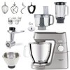 Na zdjęciu znajduje się robot kuchenny planetarny w kolorze srebrnym z logo "KENWOOD" oraz napisem "TITANIUM CHEF BAKER XL", otoczony różnymi akcesoriami, takimi jak trzepaczki, miski, malakser, blender, młynek i wyciskarka do cytrusów. Widoczne są także 
