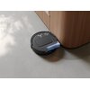 Czarny robot sprzątający porusza się wzdłuż drewnianej szafki na jasnej podłodze, z widocznym efektem działania szczotki. Na górnej części urządzenia znajduje się logo w kształcie litery "Y".