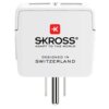 Biały adapter podróżny z widocznym logo "SKROSS" oraz napisem "ADAPT TO THE WORLD" i "DESIGNED IN SWITZERLAND". Adapter ma prostokątny kształt z metalowymi bolcami do podłączenia.