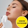 Na zdjęciu widoczna jest osoba używająca białej szczoteczki do twarzy, a w prawym dolnym rogu znajduje się żółte koło z napisem "SONICZNE OCZYSZCZENIE". Szczoteczka ma białe włosie i jest trzymana przy podbródku.