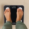 Czarna waga łazienkowa z wyświetlaczem pokazującym "52.05 kg" oraz logo "ALPHA" na dole. Na wadze stoją stopy osoby w zielonych spodniach.