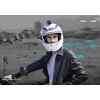 Na zdjęciu znajduje się osoba w białym kasku motocyklowym z zamontowaną kamerą, ubrana w czarną skórzaną kurtkę, siedząca na motocyklu. W tle widoczne są rozmyte postacie oraz cyfrowe efekty graficzne.