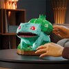 Na zdjęciu znajduje się model 3D przedstawiający postać przypominającą Bulbasaura z serii Pokemon, złożony z wielu elementów, ustawiony na okrągłej podstawie z imitacją roślinności. W tle widoczne są elementy wnętrza, takie jak półka z książkami i lampa, 