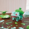 Na zdjęciu znajduje się złożony model 3D przedstawiający postać przypominającą Bulbasaura, wykonany z papierowych elementów w różnych odcieniach zieleni. Obok modelu leżą rozłożone części puzzli oraz podstawka z roślinnymi elementami.