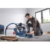 Na zdjęciu znajduje się pilarka ukosowa BOSCH Professional GCM 340-305 D, ustawiona na podłodze w jasnym pomieszczeniu z dużym oknem. Obok pilarki leżą drewniane deski, a w tle widoczny jest odkurzacz przemysłowy z podłączonym wężem.