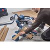 Na zdjęciu widoczna jest pilarka ukosowa BOSCH Professional GCM 340-305 D, używana do cięcia drewnianych desek. W tle znajdują się inne narzędzia i materiały budowlane.