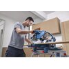 Na zdjęciu znajduje się pilarka ukosowa BOSCH Professional GCM 340-305 D ustawiona na stole roboczym, z widocznym logo BOSCH na urządzeniu. W tle znajdują się szafki kuchenne w jasnym kolorze.