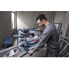 Na zdjęciu znajduje się pilarka ukosowa BOSCH Professional GCM 340-305 D, używana przez osobę w ochronnych okularach w warsztacie. W tle widoczne są metalowe ramy i szare ściany, a przez okno wpada naturalne światło.