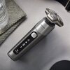 Na zdjęciu znajduje się golarka w kolorze srebrnym z trzema okrągłymi głowicami tnącymi, na której widoczny jest napis "PHILIPS 6000 Series" oraz "SkinIQ". Obok golarki leży szklanka z wodą na szarej tkaninie.
