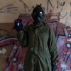 Osoba ubrana w zielony kombinezon i maskę ochronną trzyma w ręku telefon z wyświetlonym symbolem tarczy, co sugeruje ochronę urządzenia. W tle widać ścianę pokrytą graffiti, co nadaje scenie industrialny charakter.