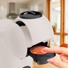Na zdjęciu znajduje się biały ekspres do kawy z widocznym miejscem na kapsułkę, w którym umieszczona jest kapsułka z napisem "Nescafé Dolce Gusto". W tle widać rozmyty obraz wnętrza kuchni z dużymi oknami.