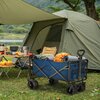 Na zdjęciu znajduje się niebieski wózek turystyczny z logo "NILS CAMP", wypełniony różnymi przedmiotami, w tym zrolowaną matą i torbą. W tle widoczny jest zielony namiot oraz stół kempingowy z naczyniami i owocami, a całość znajduje się na trawiastej powi