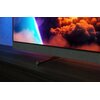 Telewizor z cienką ramką wyświetla kolorowy obraz z chmurami, a jego podstawa ma napis "Philips". Urządzenie stoi na ciemnym drewnianym blacie, a w tle widać niebieską ścianę.