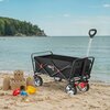 Czarny wózek turystyczny z regulowaną rączką stoi na piaszczystej plaży, obok zamku z piasku i kolorowych zabawek. Na boku wózka widoczne jest logo "NILS".