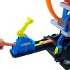Zestaw toru wyścigowego Hot Wheels Mega Garaż Gigapętla z widocznym niebieskim elementem z logo Hot Wheels i żółtym mechanizmem, na którym spoczywa palec. W tle znajduje się pomarańczowa pętla oraz różnokolorowe elementy toru.