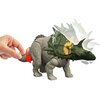 Na zdjęciu znajduje się szara figurka dinozaura z zielonymi i pomarańczowymi elementami na głowie, której ogon jest pociągany przez rękę, co powoduje ruch głowy. W tle widoczne są rozmyte kontury głowy dinozaura, sugerujące jej ruch.